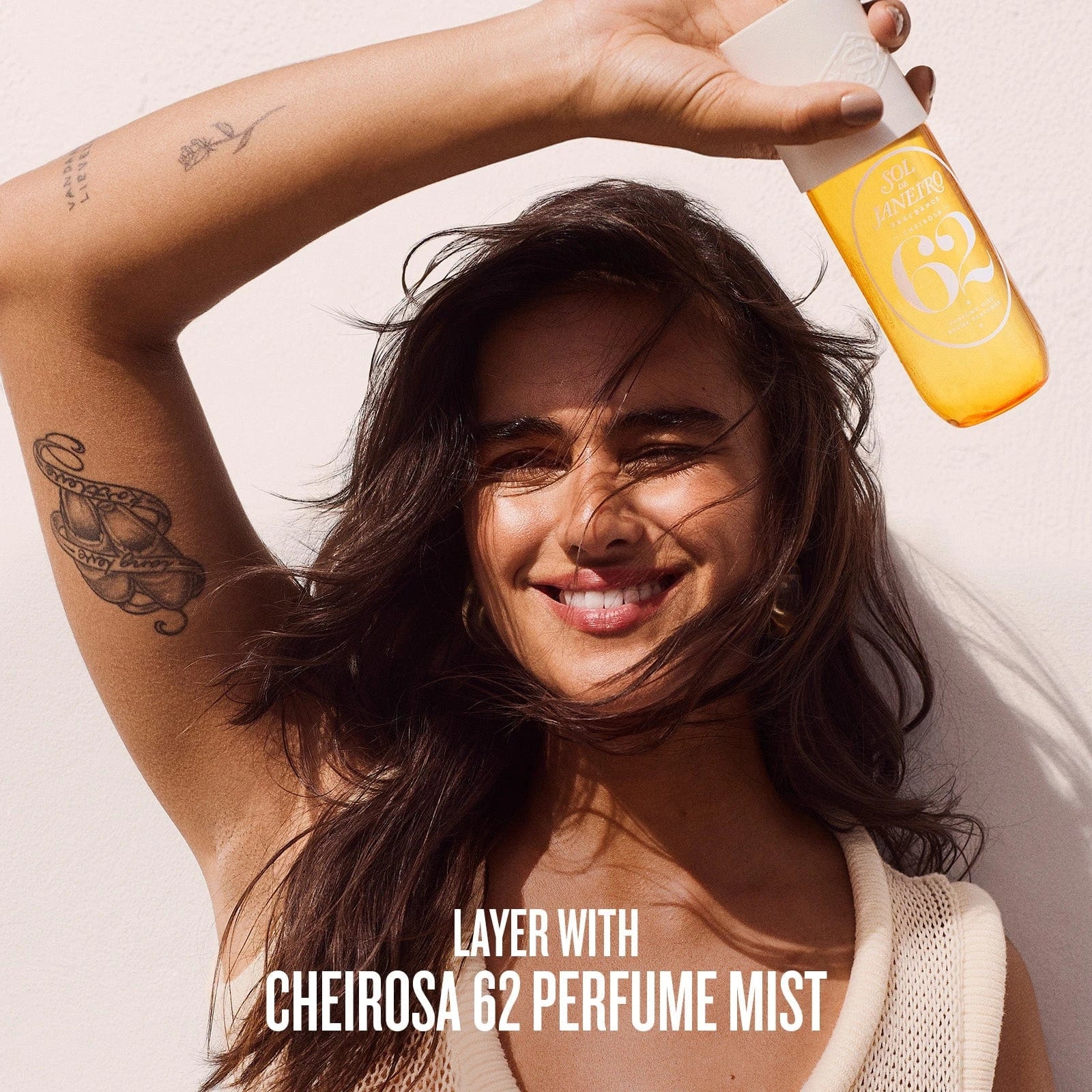 This image is of model smiling while holding Sol de Janeiro Cheirosa 62 Perfume Mist in her hand. The text on the image is as follows "layer with cheirosa 62 perfume mist"