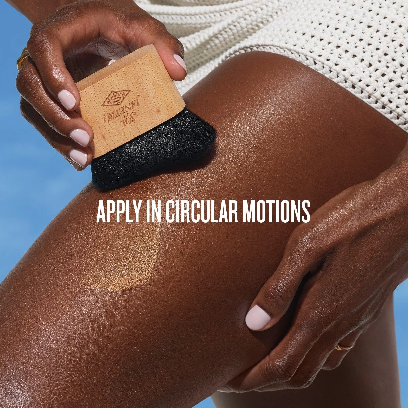 text on the image reads apply in circular motions. This image shows model using the brush to put on her sol de janeiro glowmotions body oil on her legs