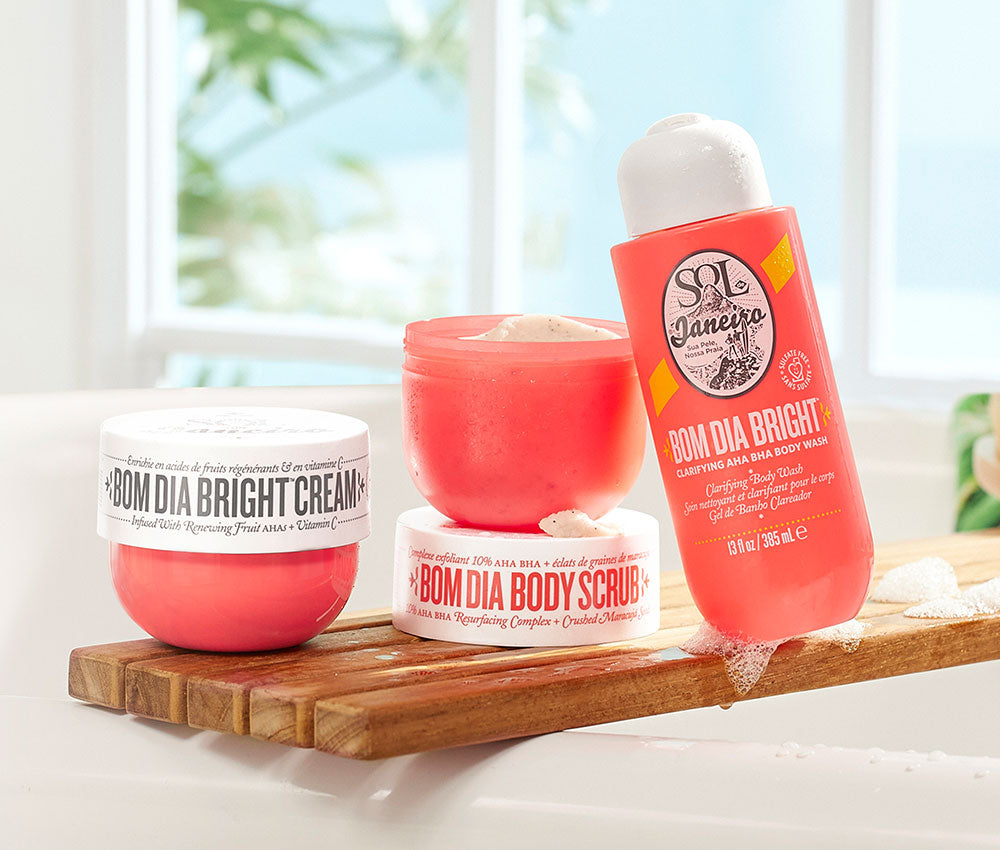Bom dia bright body cream, bom dia body scrub, and bom dia body wash sitting on a wooden shelf across a bathtub
